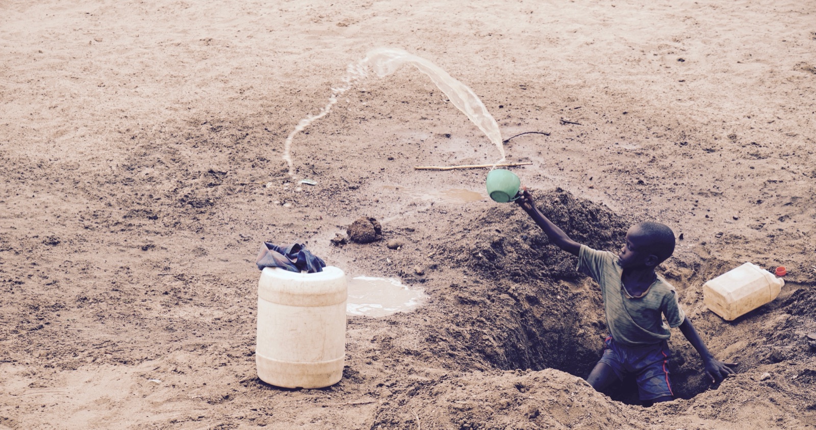 A boy in a shallow well pours water into a plastic drum, Kakuma, Kenya
